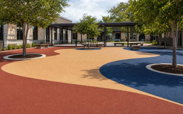 A school play courtyard in red and tan rubber surfacing under shade trees.